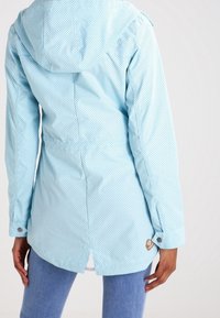 Ljusblå vattentät jacka med huva, med ett prickmönster, tryckknappsstängning vid ärmarna och en lätt svans för extra skydd.