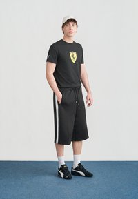 Νέος άνδρας με μαύρο t-shirt Ferrari, μαύρο σορτς με λευκές ρίγες, μαύρα αθλητικά παπούτσια Puma, λευκές κάλτσες και λευκό καπέλο, στέκεται σε μπλε πατώμα.