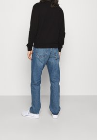 Una persona in piedi con le spalle verso la fotocamera, che indossa un felpa nera, jeans blu e sneakers bianche, su uno sfondo semplice chiaro.