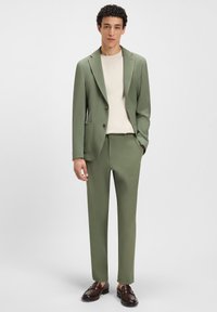 Lichtgroend pak van gestructureerde stof met twee knopen en voorzakken, gecombineerd met een crèmekleurig overhemd en bruine loafers.
