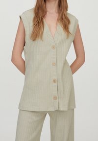 Mujer vistiendo un chaleco sin mangas de canalé beige con botones y pantalones de canalé a juego, de pie con las manos en la espalda sobre un fondo plano.