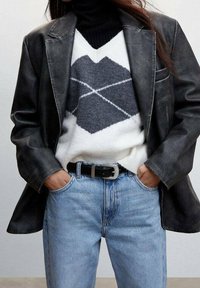 Blazer de cuero negro sobre un suéter blanco con un patrón geométrico gris, combinado con jeans de un azul claro y un cinturón negro.