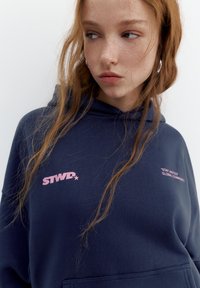 Giovane donna con lunghi capelli rossi che indossa una felpa con cappuccio blu navy con testo rosa "STWR" e "Stay United Global Community."