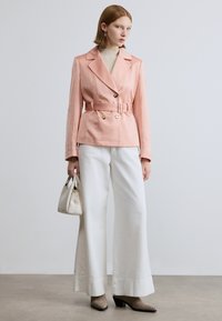 Διπλόσχεδη ροδακινί ζακέτα με ζώνη στη μέση, συνδυασμένη με λευκό παντελόνι με φαρδιά μπατζάκια και γκρι τακούνια. Μικρή λευκή τσάντα ως αξεσουάρ.