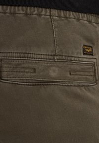 Tissu vert olive avec une poche arrière cousue, comportant un petit badge logo. La taille est noire et élastique.