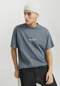 T-shirt gris à manches courtes avec un col rond et un logo blanc sur le devant, associé à un jean en denim foncé et à un bonnet en tricot léger.