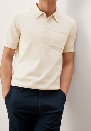 Man draagt een crème korte mouw polo met een zakje, ingestopt in donker marineblauwe broek, handen in de zakken, staand tegen een lichte muur.