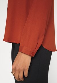Chemise à manches longues de couleur rouge profond, réalisée en tissu léger. Elle présente des poignets boutonnés et un ourlet incurvé, portée avec un pantalon noir ajusté.