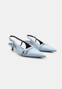 Talons slingback en cuir bleu clair avec des pointes, présentant deux sangles avec des boucles en argent et un petit talon carré. Texture lisse.