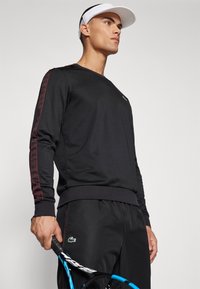 Sudadera negra de manga larga con patrones de rayas rojas a lo largo de las mangas, con un pequeño logo verde y una tela texturizada.