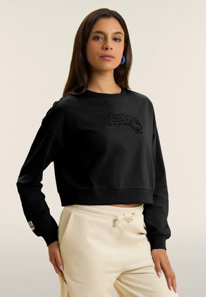 Woman wearing a black cropped sweatshirt with "FREDDY" text and graphic, paired with beige drawstring pants, blue hoop earrings.