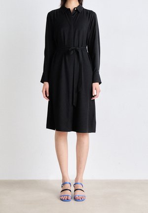 Schwarzes Hemdblusenkleid mit Kragen, langen Ärmeln, gebundener Taille und Knopfleiste vorne. Kombiniert mit blauen und schwarzen Riemchensandalen. Glatte Stoffstruktur.