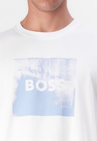 Fehér pamut póló, kék téglalap alakú grafikával, amelyen a "BOSS" szó vastag fehér betűkkel szerepel, textúrált felülettel.