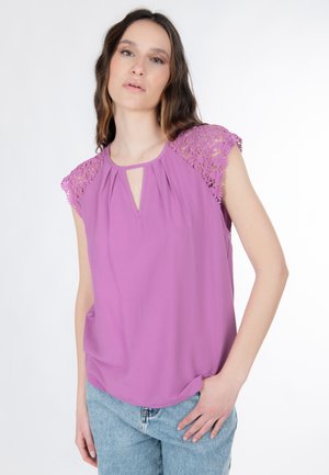 Donna con indosso una blusa lavanda senza maniche con maniche ad aletta in pizzo e scollo a goccia, abbinata a jeans azzurri, in piedi con una mano in tasca.