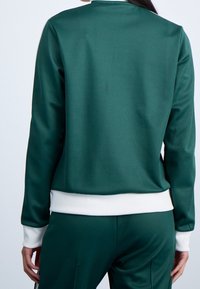 Person i en mörkgrön träningsoverall med vita manschetter och midja, sedd bakifrån mot en neutral bakgrund.