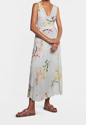 Frau trägt ein ärmelloses weißes Maxikleid mit mehrfarbigen Blumenmustern und roten flachen Sandalen mit Riemen.
