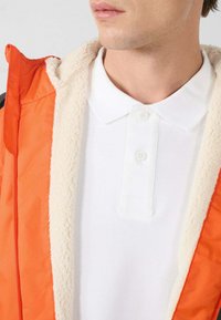 Oranje jas met ritssluiting en een zachte witte fleece voering, gedragen over een effen wit poloshirt met een button-down kraag.