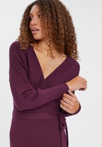 Robe à manches longues en tissu côtelé violet foncé avec un décolleté en V et une ceinture à la taille, présentant des détails texturés et des poignets de manches bi-ton.