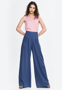 Różowy top bez rękawów z dekoltem w kształcie litery V zestawiony z wysokim stanem szerokimi spodniami w kolorze granatowym z plisami. Uzupełniony wzorzystymi butami na platformie.