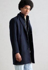 Abrigo de lana navy con cuello con muesca, tres botones y bolsillos laterales sobre una chaqueta negra con cremallera. Llevado con jeans de color azul claro.
