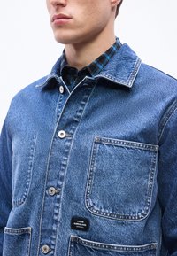 Άνδρας που φορά μπλε τζιν μπουφάν με μπροστινές τσέπες πάνω από μαύρο και μπλε καρώ πουκάμισο, ορατός από τους ώμους μέχρι το πηγούνι.