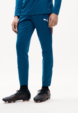 Parte inferior del cuerpo de una persona que lleva pantalones deportivos azules Puma y botas de fútbol negras Puma con detalles naranjas sobre fondo blanco.