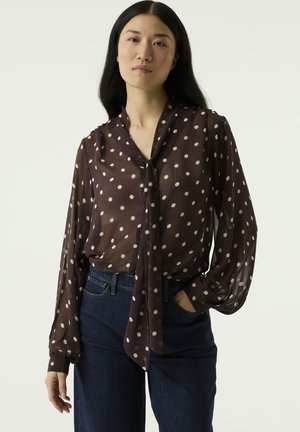 Vrouw met lang zwart haar die een doorschijnende bruine blouse met witte stippen en donkerblauwe spijkerbroek draagt, één hand in de zak, neutrale gezichtsuitdrukking.