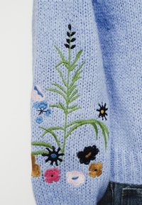 Un pull en tricot bleu clair avec une broderie florale complexe en vert, rose, bleu, noir et jaune sur la manche gauche.