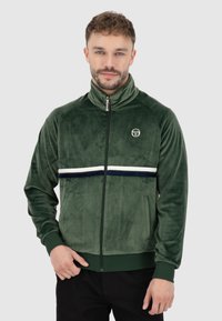Giacca in velour verde con zip, colletto alto, striscia orizzontale bianca e blu, tasche con zip e logo sul petto.