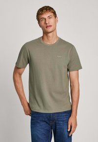 T-shirt a maniche corte verde oliva in cotone, con colletto rotondo e piccolo dettaglio del logo sul lato sinistro del petto. Indossato con jeans blu.