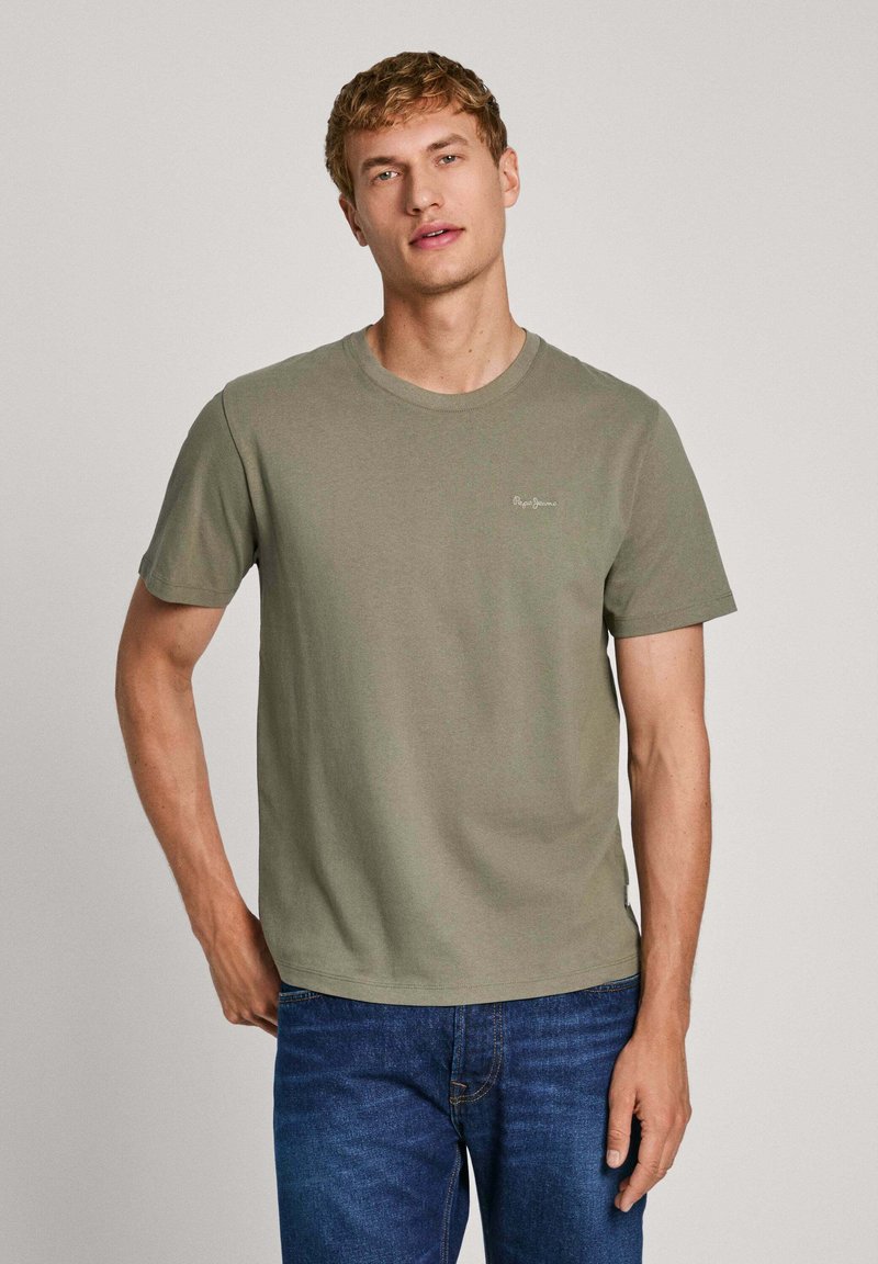 T-shirt a maniche corte verde oliva in cotone, con colletto rotondo e piccolo dettaglio del logo sul lato sinistro del petto. Indossato con jeans blu.