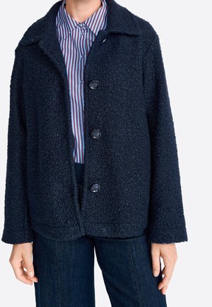 Person iført en tekstureret mørk marineblå knapjakke over en blå og mørkerød stribet skjorte med krave og mørkeblå jeans.