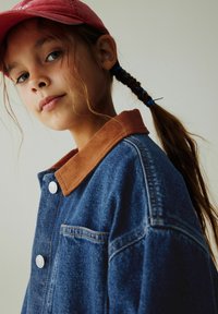 Džinsinė striukė su rudos spalvos kordurojo apykakle, sidabriniais mygtukais, priekinė kišene ir laisvo kirpimo, dėvima su raudonu kepuru.