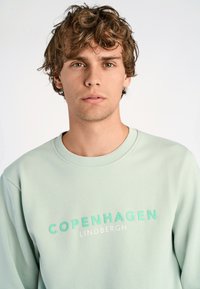 Vaaleanvihreä collegepaita, jossa on pyöreä kaula-aukko. Edessä rohkea teksti "KOPENHAGEN" mintunvärisenä ja "LINDBERGH" valkoisena. Pehmeä kangas, rento malli.