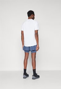Camiseta atlética de manga corta blanca con mangas raglán, combinada con pantalones cortos de color azul oscuro. Zapatillas y calcetines negros completan el atuendo.