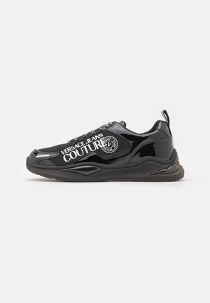 Schwarzer Sneaker in niedriger Ausführung mit glänzendem Finish, klobiger Sohle, dem Schriftzug "VERSACE JEANS COUTURE" und einem runden Logo an der Seite.