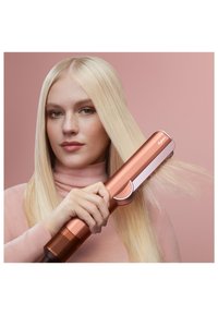 Piastra per capelli Dyson in oro rosa con design liscio ed elegante e impugnatura testurizzata, tenuta da una persona con capelli biondi, lunghi e lisci.