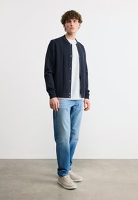 Νέος άνδρας σε όρθια θέση, φορώντας ένα ναυτικό μπλε σακάκι με κουμπιά πάνω από ένα λευκό πουκάμισο, ανοιχτόχρωμο τζιν και μπεζ αθλητικά παπούτσια σε γκρι πάτωμα.