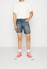 Persona que lleva una camiseta blanca, pantalones cortos de mezclilla de lavado medio con bordes deshilachados, calcetines blancos y zapatillas de caña alta color coral, contra un fondo liso.