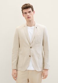 Blazer en lin beige avec un devant à bouton simple, poche à rabat et deux poches latérales. Porté par-dessus un T-shirt blanc, présentant une texture lisse.