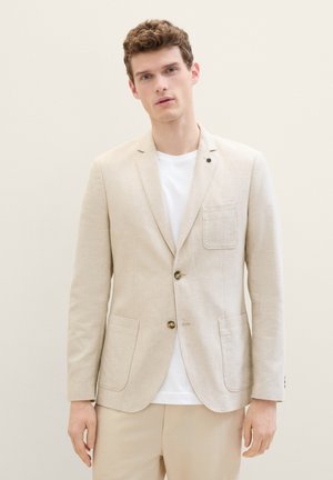 Blazer en lin beige avec un devant à bouton simple, poche à rabat et deux poches latérales. Porté par-dessus un T-shirt blanc, présentant une texture lisse.