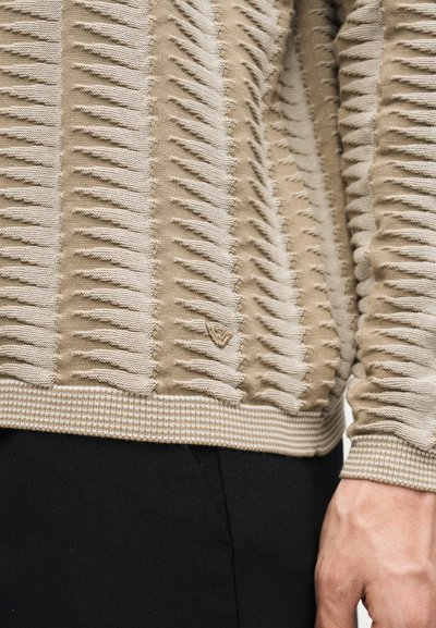 Pull en maille texturée beige et blanc avec ourlet et poignets côtelés, porté avec un pantalon noir, montrant un gros plan du torse et de la main droite.