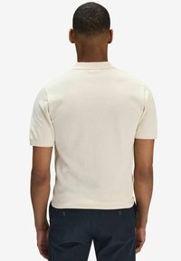 Kortärmad polo i ljus krämfärg, med texturerad tyg. Klassisk krage och rak nederkant. Baksidan visas.