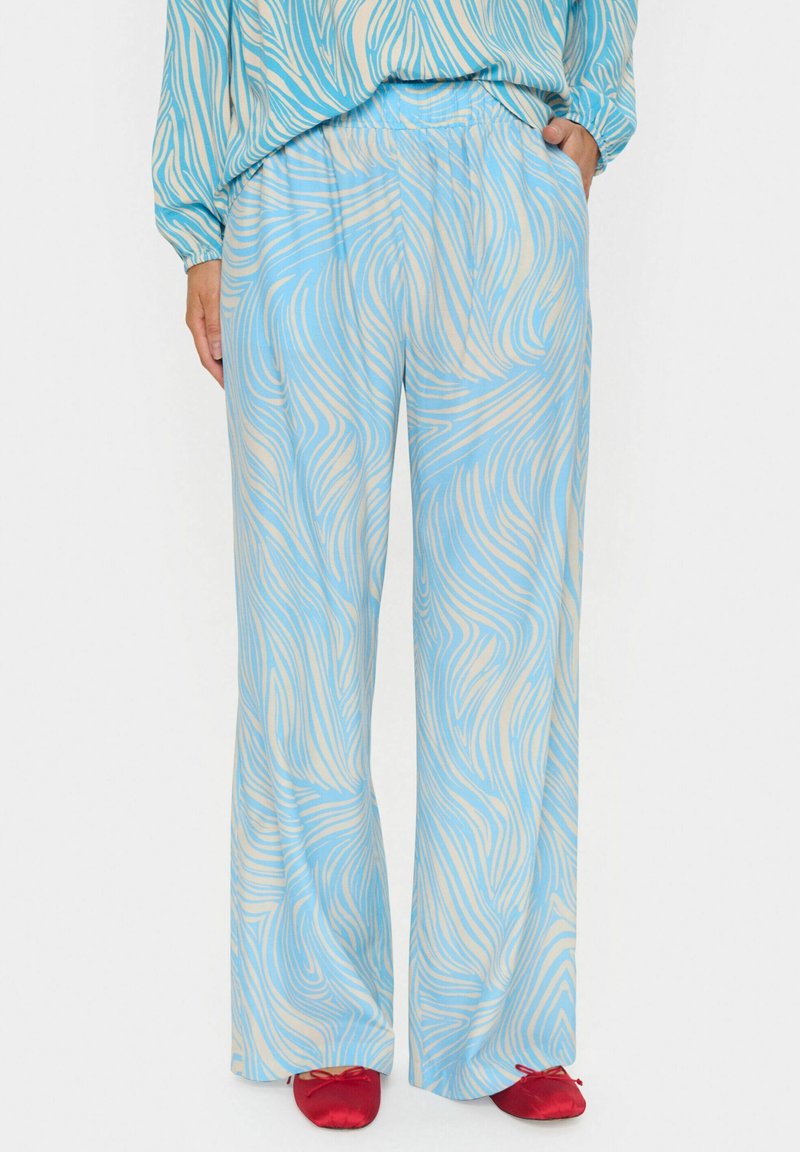 Pantalon large à motif abstrait tourbillonnant bleu clair et beige, porté avec un haut assorti et des ballerines rouges, main dans la poche.