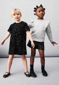 Černé sametové šaty s bílými puntíky; šedý svetr s volánkovými detaily; černé kožené šortky; černé boty s mašlemi.