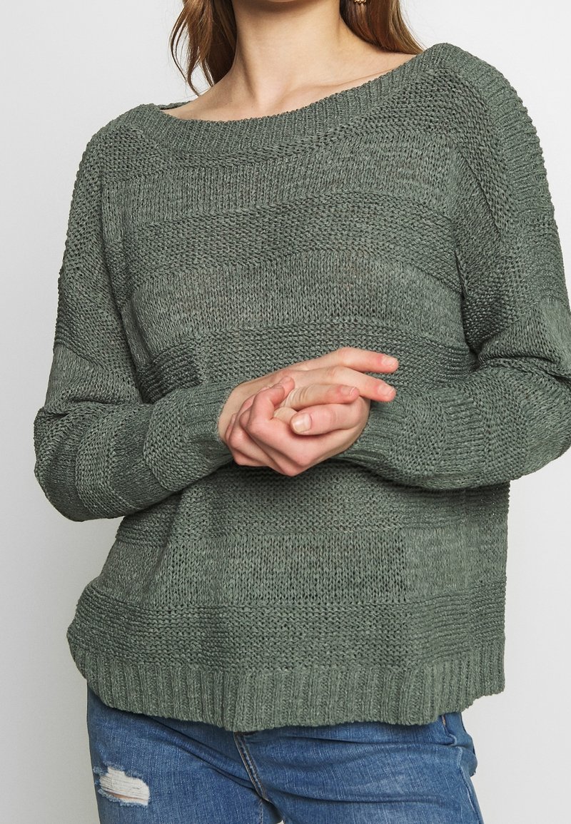 Femme portant un pull en maille texturée vert et un jean bleu déchiré, les mains doucement croisées devant elle.