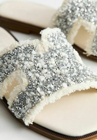Platta sandaler med en beige fusk läder fotbädd och ett dekorativt band med silver- och vita juveler, strukturerad fransdetaljering och en slät sula.