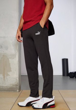 Person trägt schwarze Puma-Sweatpants, weiße Puma-Sneaker und ein rotes Shirt, steht drinnen in der Nähe von Fitnessgeräten.