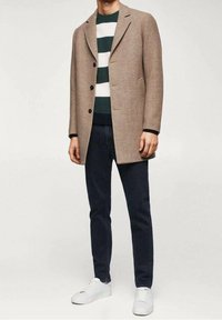 Brązowy płaszcz z mieszanki wełny z klapowanym kołnierzem, dwoma guzikami i bocznymi kieszeniami, noszony na zielono-białym paskowanym swetrze, ciemnych dżinsach i białych trampkach.