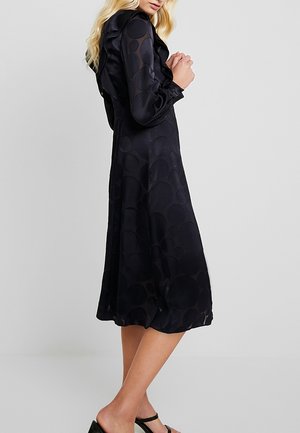 Femme portant une robe midi noire à manches longues avec de grands cercles texturés et des détails à volants, associée à des talons ouverts noirs.